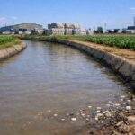 トルコの農業地帯を流れる灌漑用水路とその近くにあるプラスチックリサイクル施設を写した写実的な風景イメージ